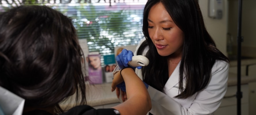 A clinician in a white coat examines a patient’s forearm with a handheld dermatoscope